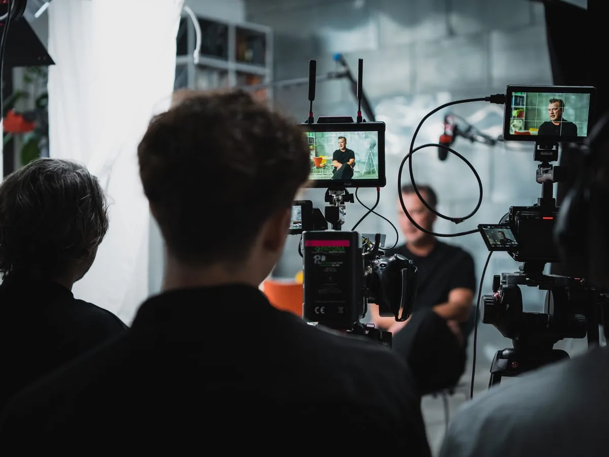 Am Set: Interview an zwei Regiemonitoren Content Creation am Set: Blick über die Schultern des Teams auf zwei Regiemonitore, auf beiden derselbe Mann im Interview, dazwischen Kamera und Angel
