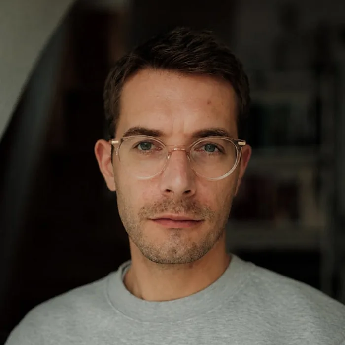 Joscha Ortmeier — Le Werk, Köln Joscha Ortmeier, Head of Post-Production bei Le Werk in Köln, mit heller Brille und grauem Sweatshirt vor einem Bücherregal